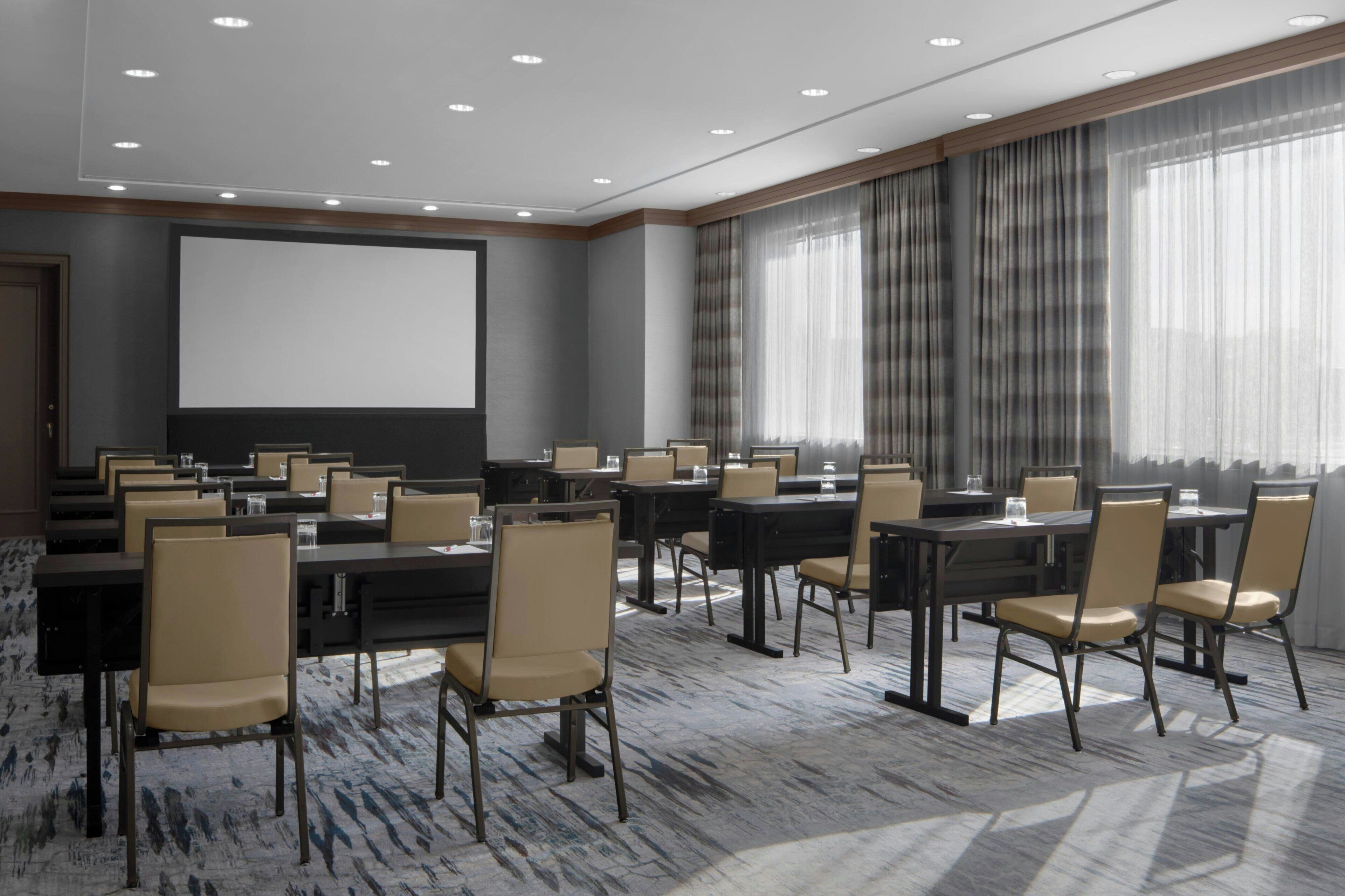 Une salle de conférence équipée de tables et de chaises, d’un écran de projection et de grandes fenêtres avec des rideaux.