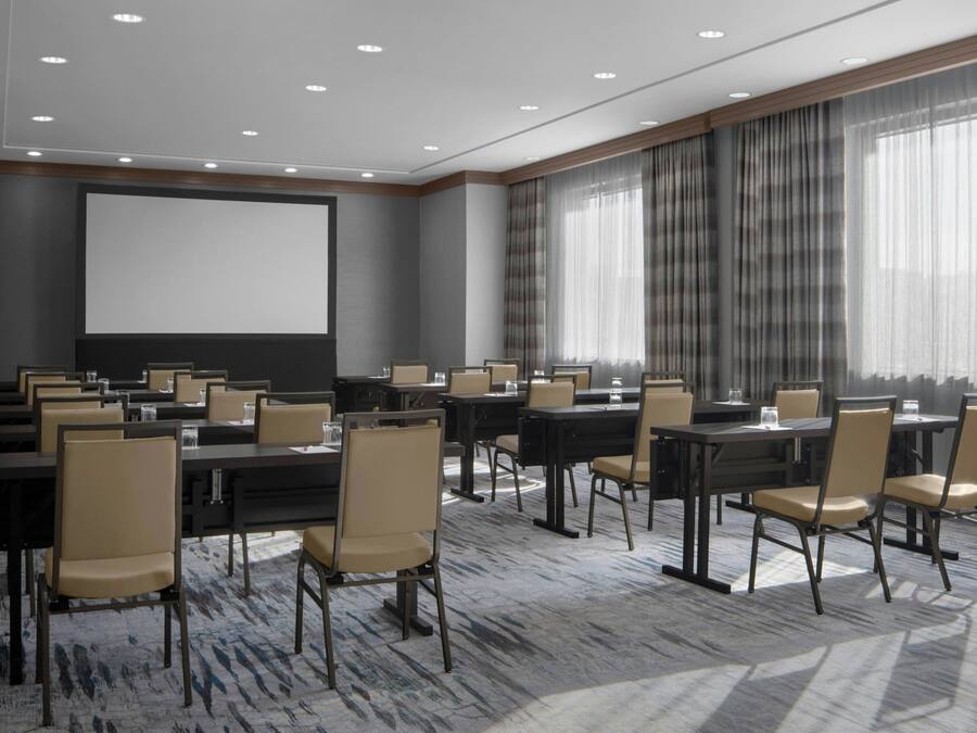 Un salón de conferencias con mesas y sillas, una pantalla de proyector y ventanas grandes con cortinas.