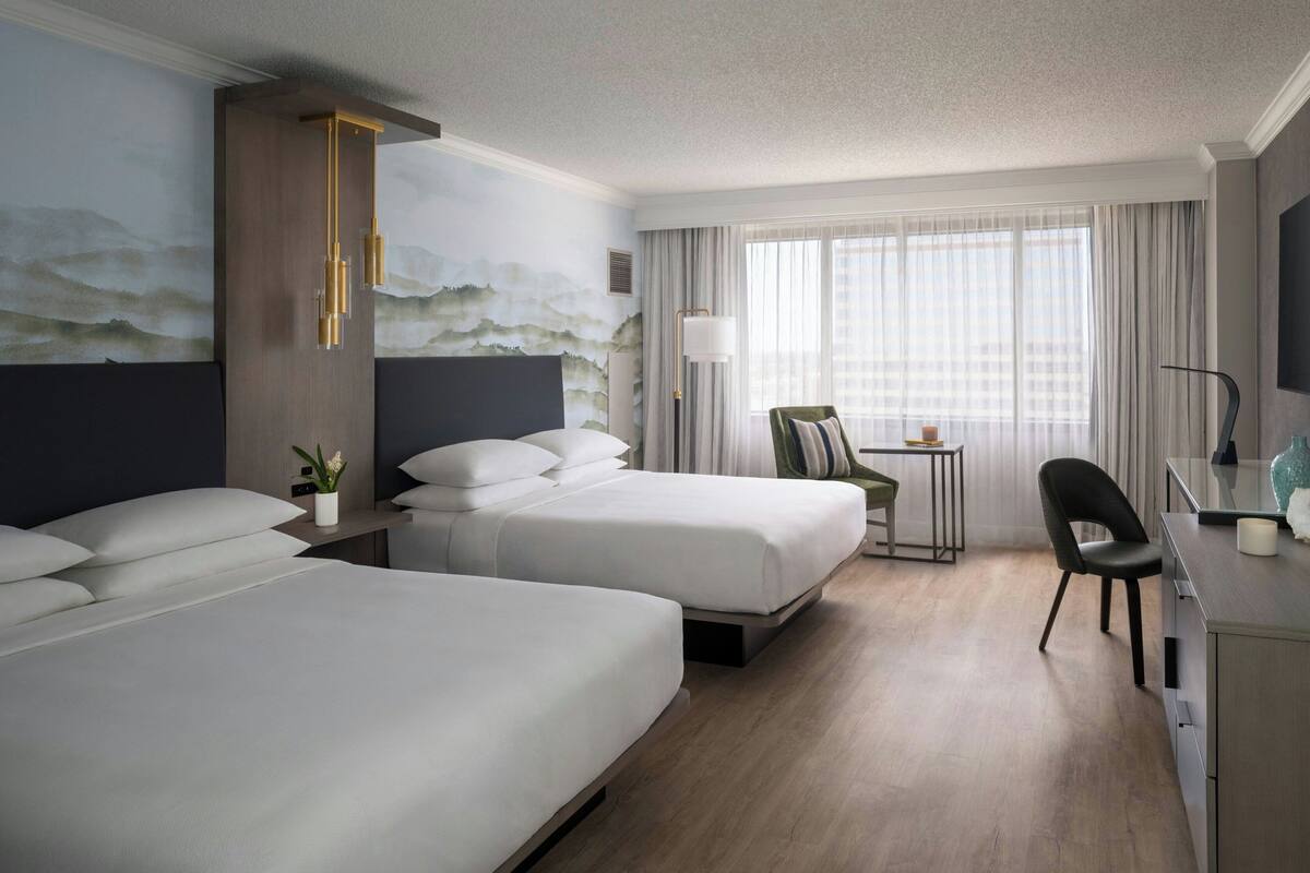 A hotel room with two beds, a desk, a chair, a lamp, and a painting on the wall.