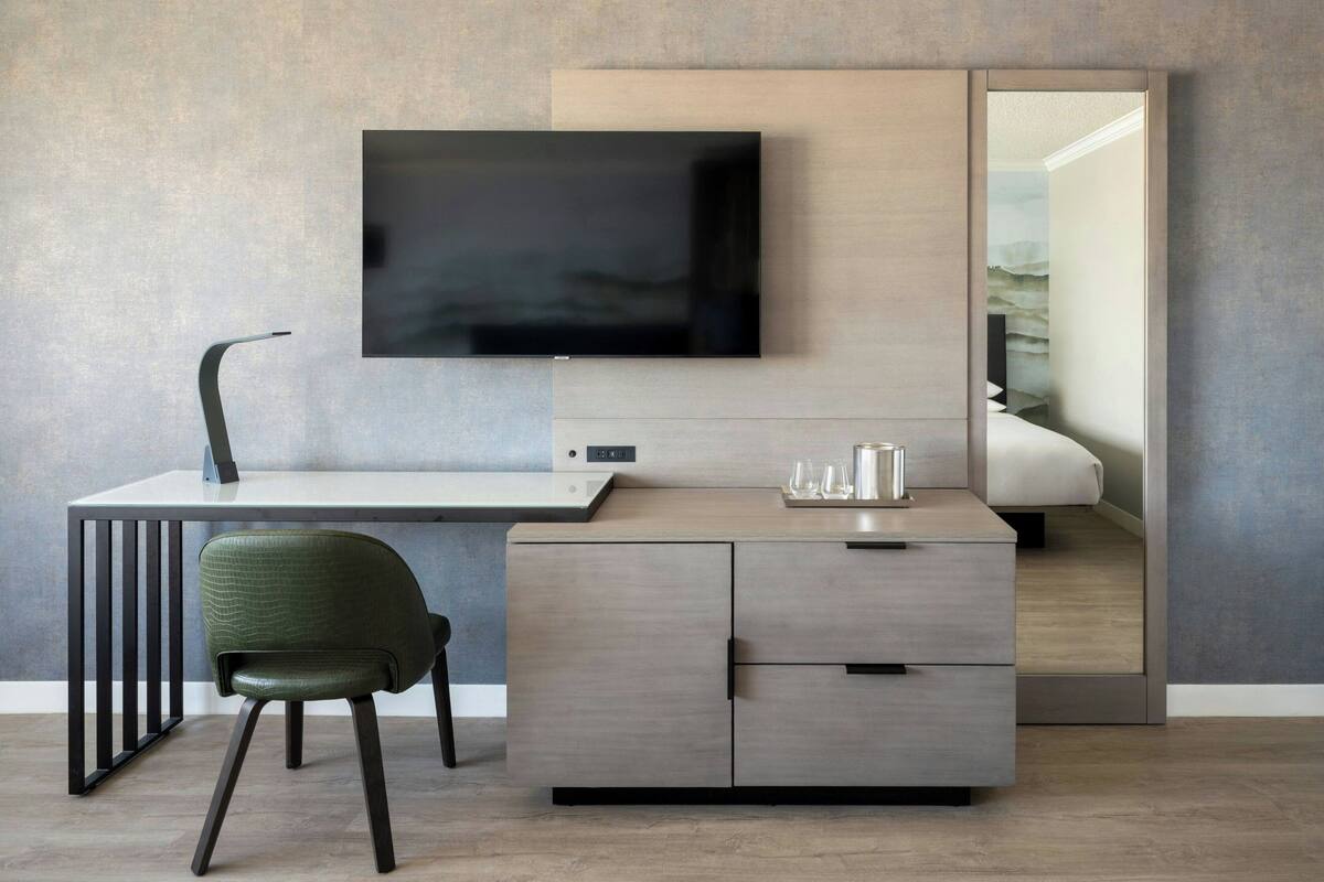 A modern hotel room with a large flat-screen TV mounted on the wall, a wooden desk with a chair, and a built-in wardrobe.