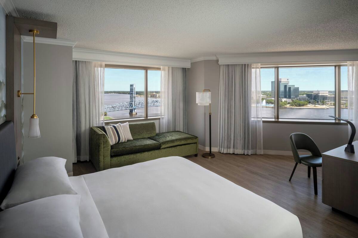 A hotel room with a large bed, a green sofa, a desk, and a view of a bridge and buildings.