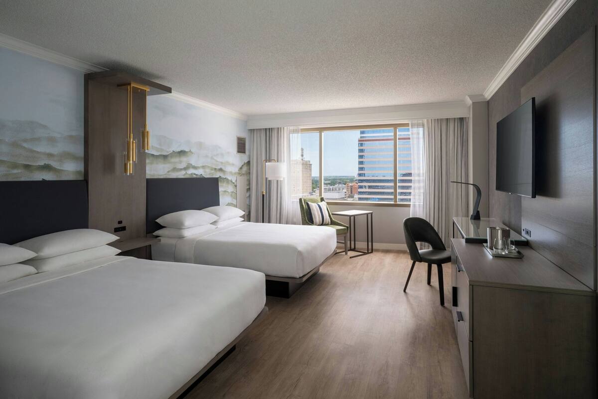 A hotel room with two beds, a desk, a chair, a TV, and a window with a city view.
