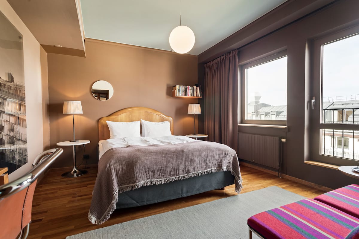 Habitación de hotel impecable con una cama grande, lámparas de noche, una mesita redonda pequeña, una estantería y un ventanal con vistas.