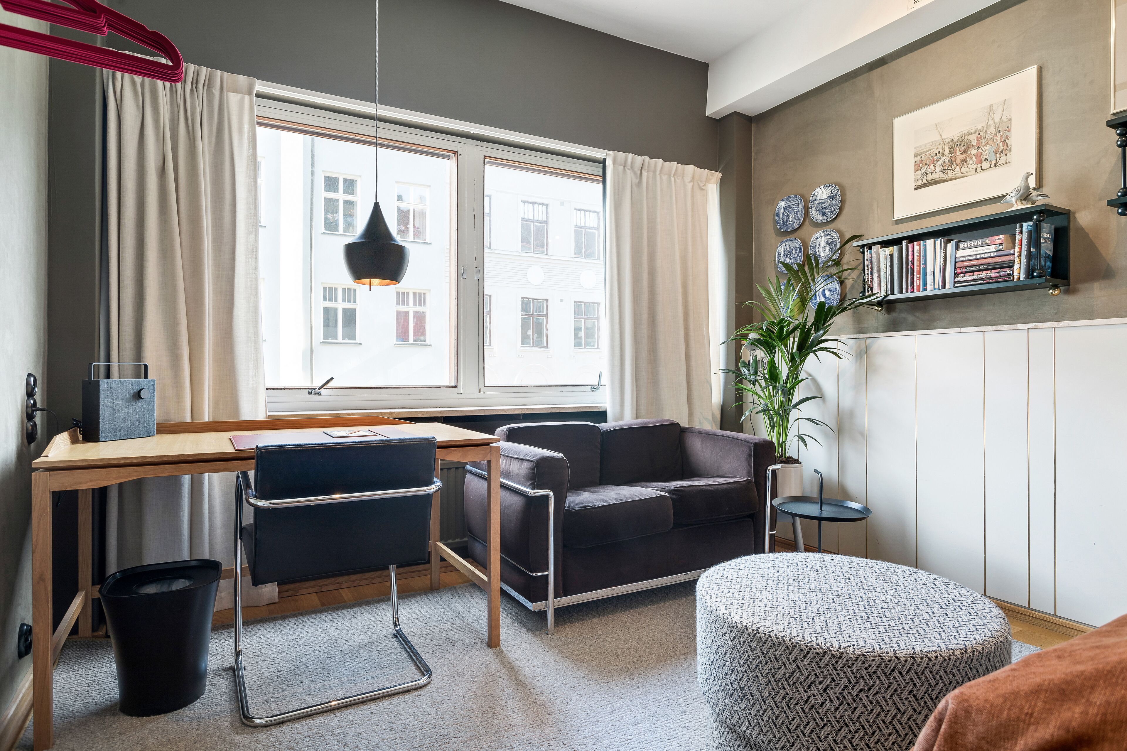 A modern living room with a wooden desk, black leather sofa, and a round ottoman. A bookshelf with books and decorative plates is on the wall.
