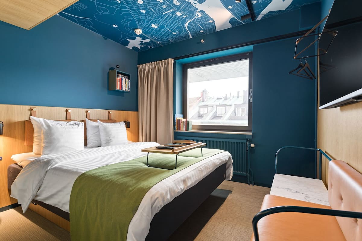 Habitación de hotel con cama, escritorio, ventana con vistas a edificios, mapa en el techo y estantería.