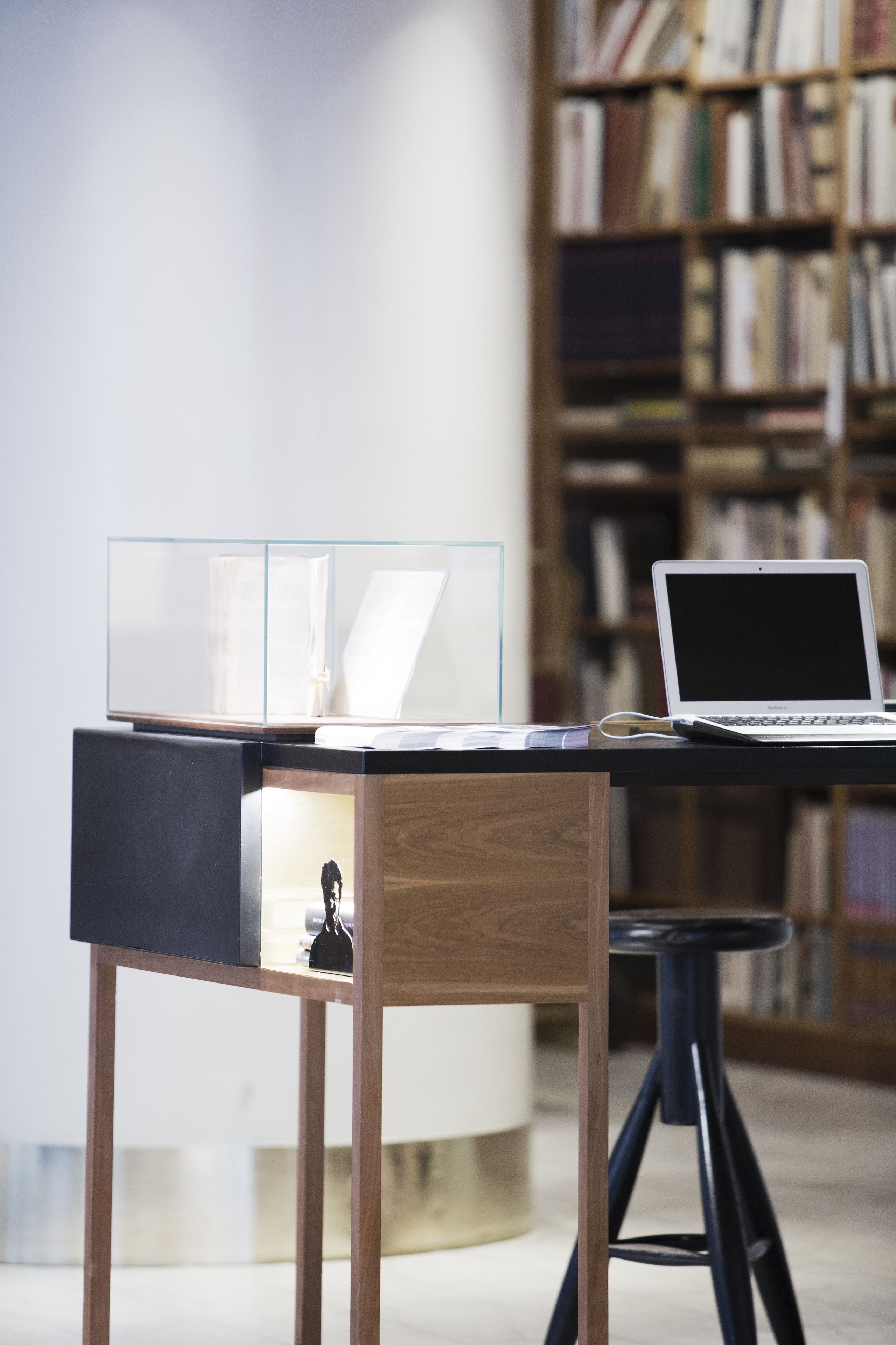 A modern workspace with a laptop on a desk, a glass display case, and a bookshelf in the background.