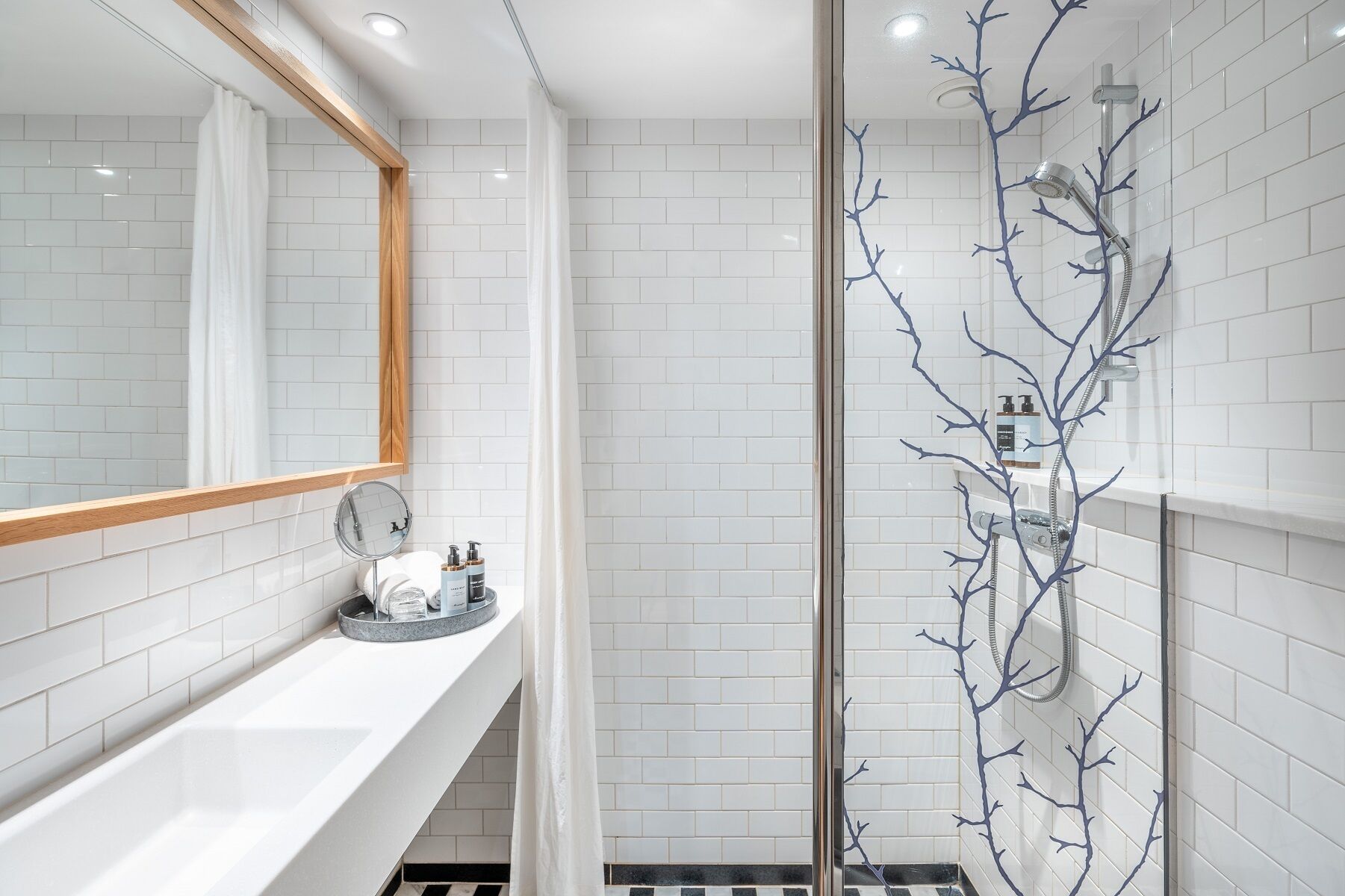 A modern bathroom with white subway tiles, a wooden-framed mirror, a glass shower enclosure with a branch-like design, and a white sink with toiletries.