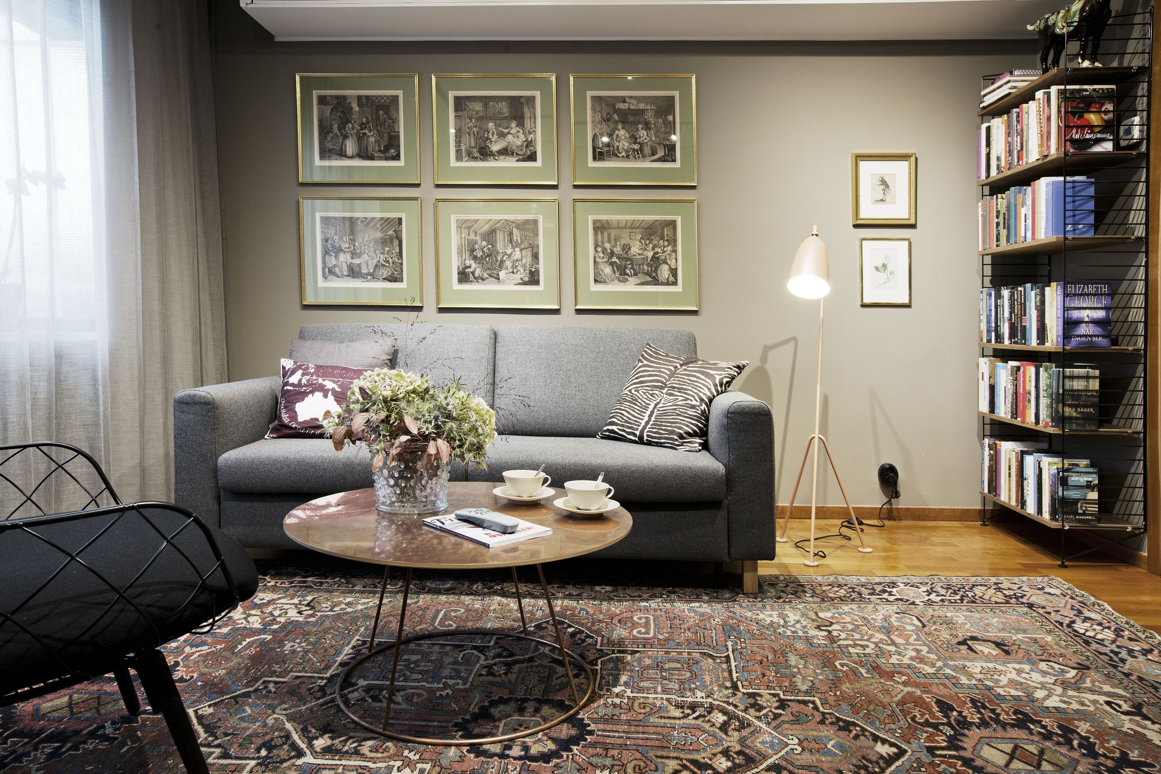A living room with a grey sofa, a coffee table with a vase and cups, a patterned rug, and a bookshelf with books.