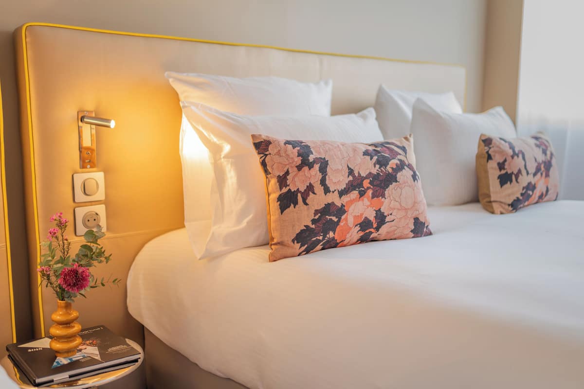A neatly made bed with white linens and floral-patterned pillows, a bedside table with a book and a vase of flowers, and a wall-mounted light fixture.