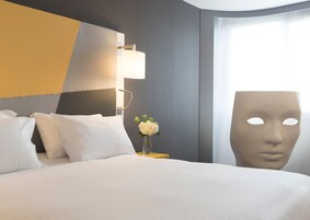 Una habitación de hotel con una cama, una mesita de noche con un florero y una lámpara de pared, además de un ventanal grande.