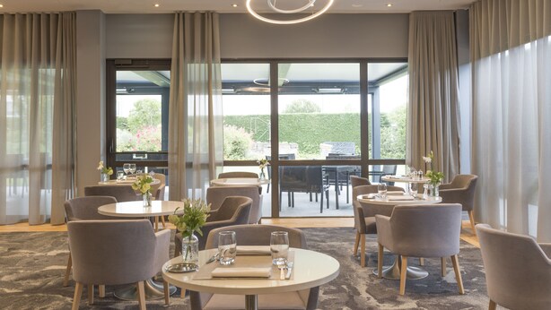 A modern dining area with round tables, comfortable chairs, and large windows offering a view of the outdoors.