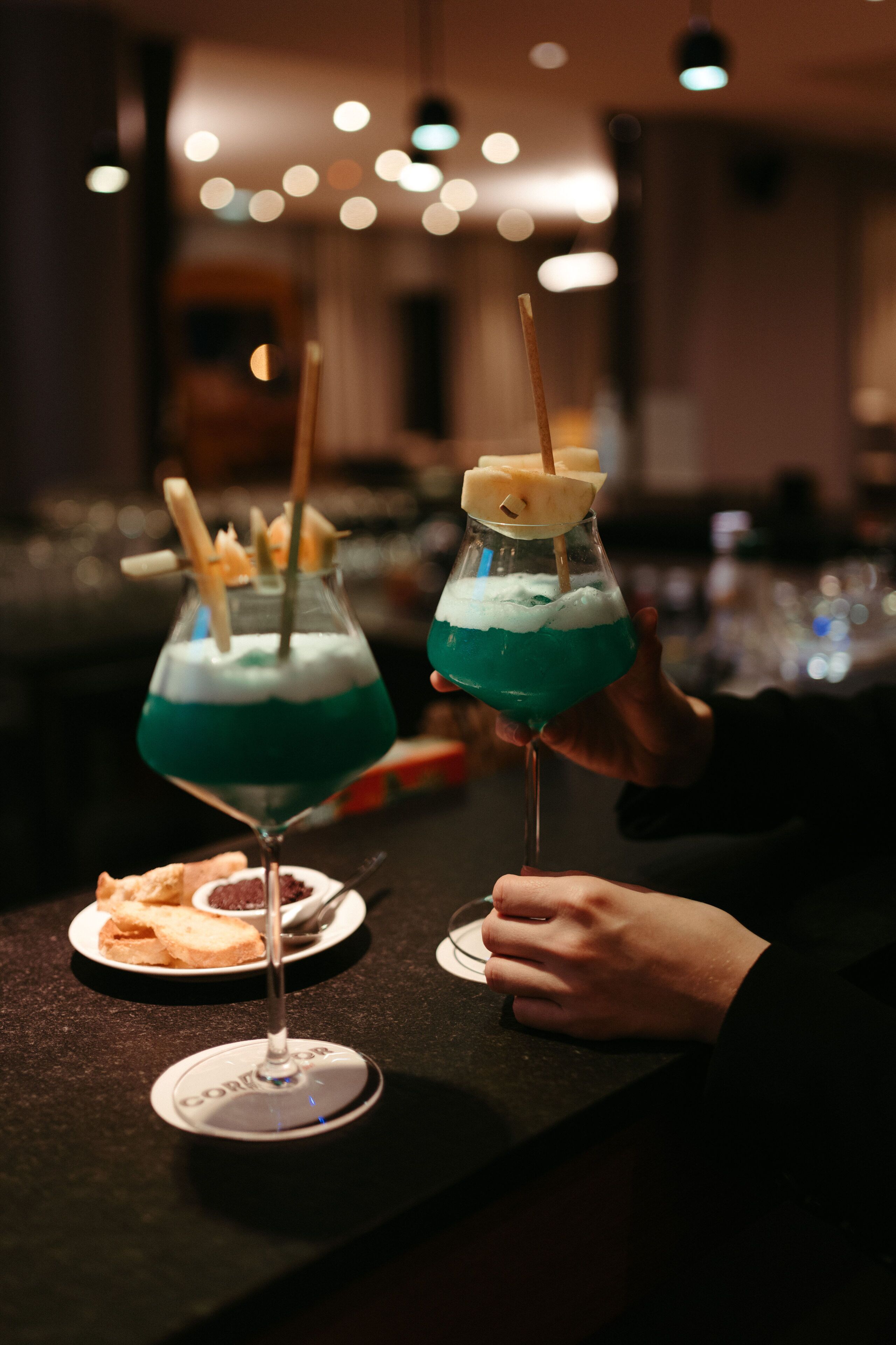 Two cocktails in glasses with straws and a slice of pineapple on the rim, placed on a bar counter with a plate of crackers.