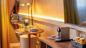 A modern hotel room with a round table, a lamp, a flat-screen TV, and a small refrigerator.