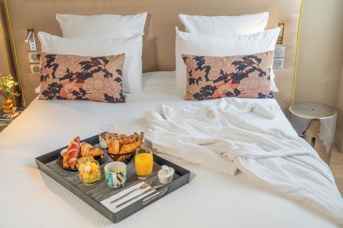 A neatly made bed with floral pillows, a tray with breakfast items, and a bedside table with a lamp.