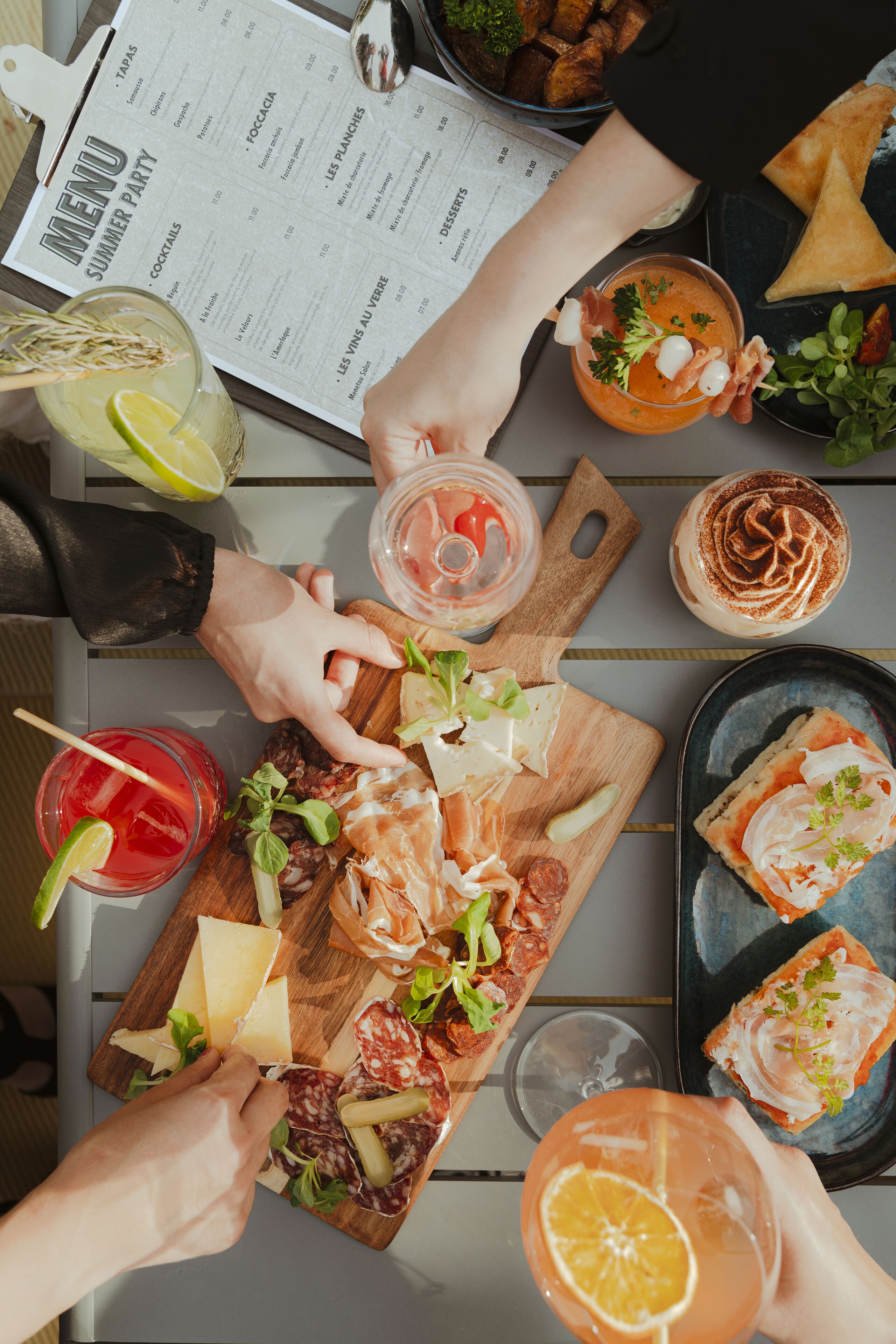 Ein Tisch mit verschiedenen Speisen und Getränken, darunter eine Speisekarte, ein Holzbrett mit Käse und Wurstwaren sowie mehrere Cocktailgläser.