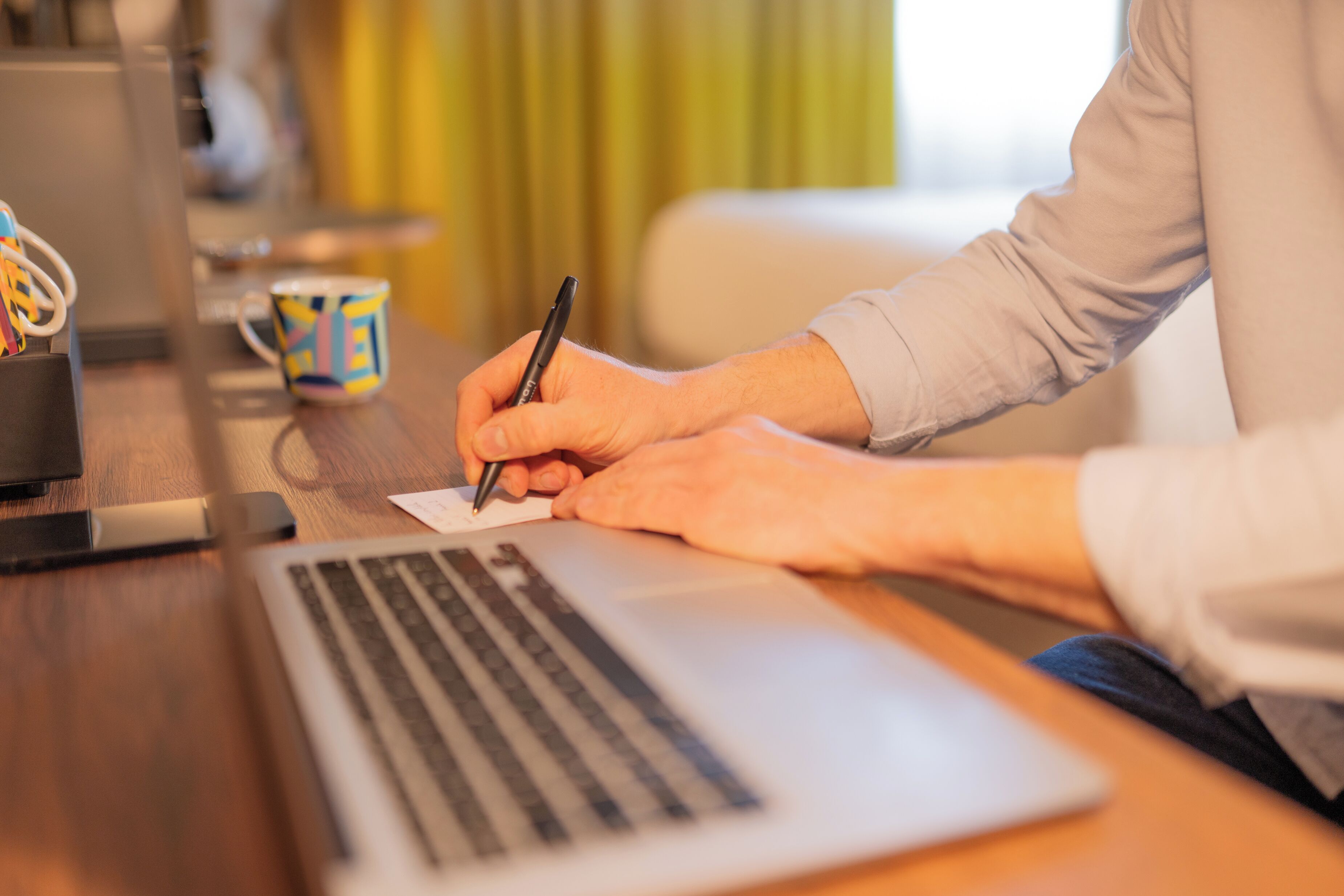 Iemand schrijft met een pen op een stuk papier, met een laptop en een mok op een houten tafel.