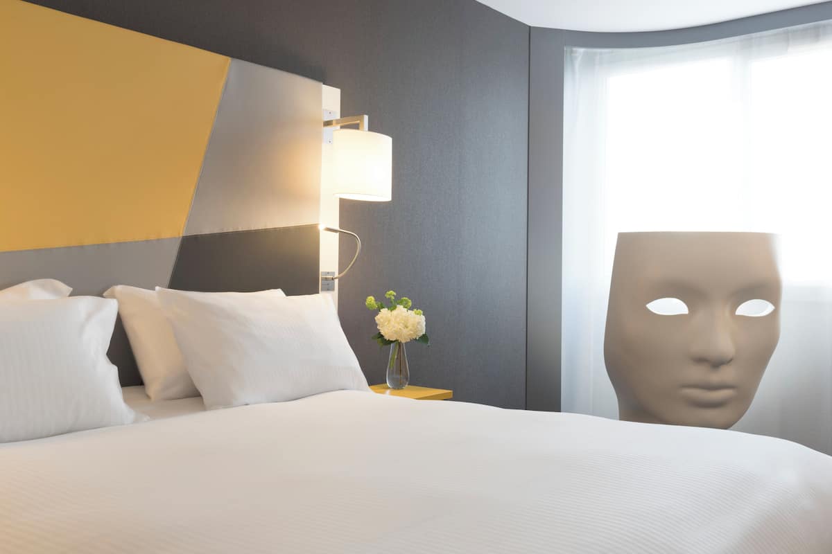 A hotel room with a bed, pillows, a nightstand, a lamp, and a vase with flowers.