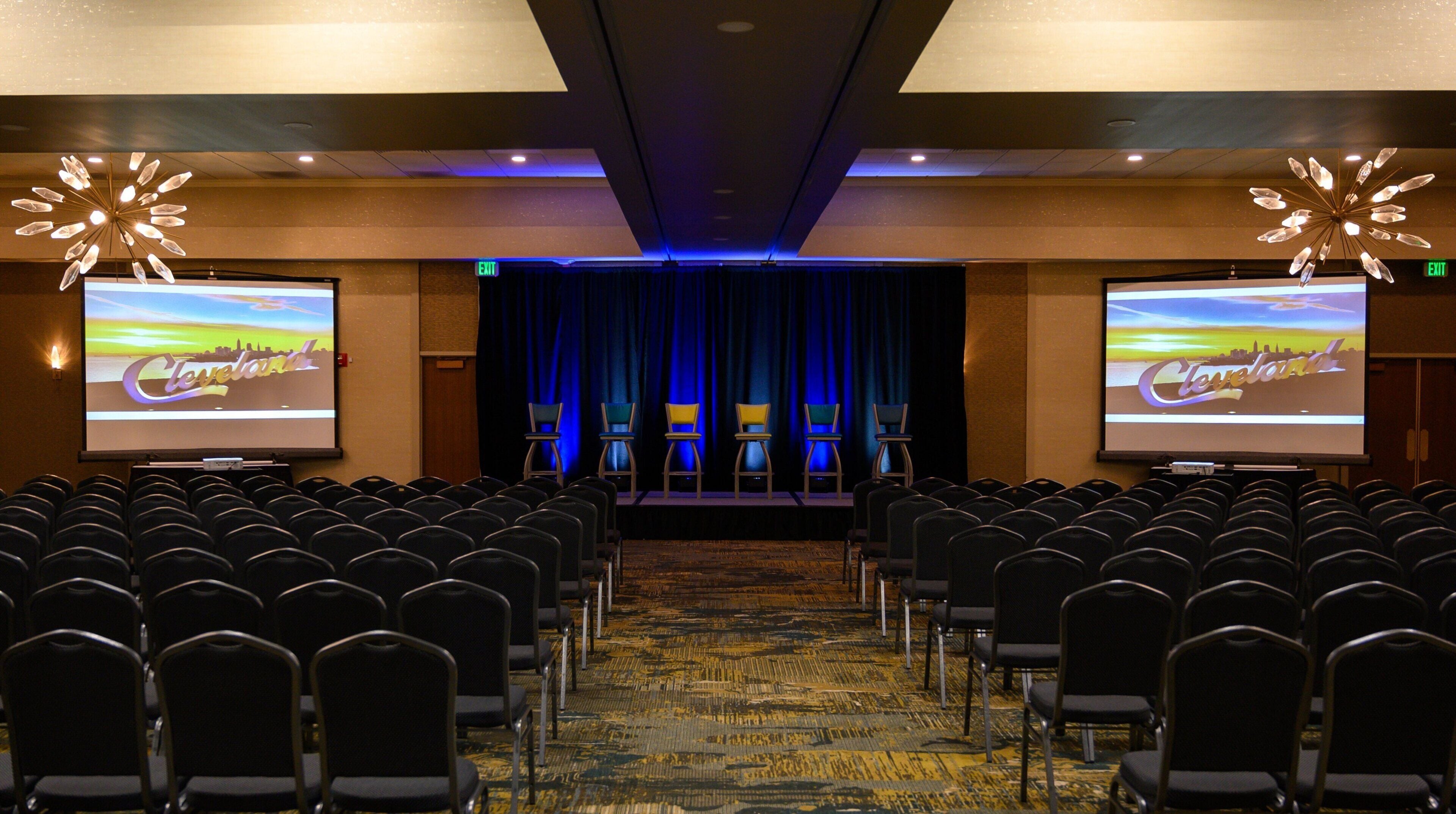 A conference room with rows of chairs, a stage with a blue curtain, and two screens displaying the word 'Cleveland'.