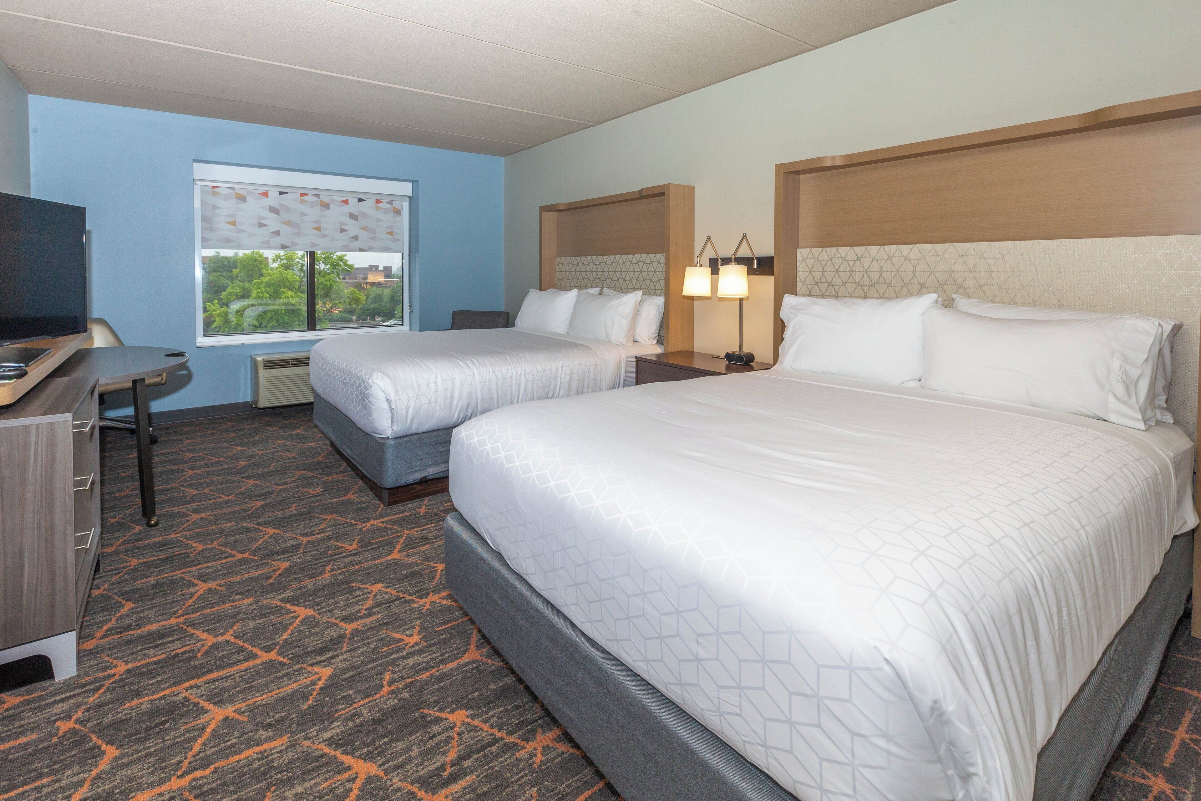 A hotel room with two beds, a desk with a TV, and a window with blinds.