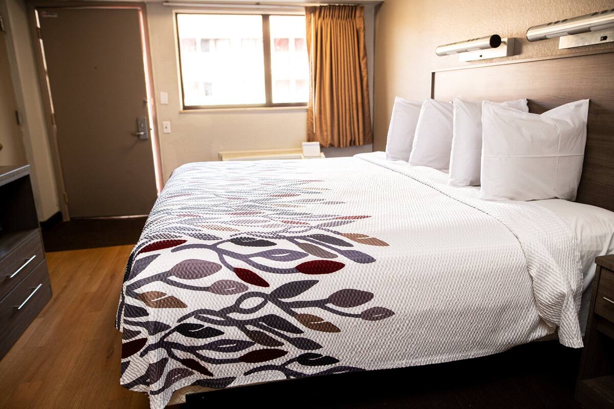 A hotel room with a bed, a dresser, a window with curtains, and a headboard with a light fixture.