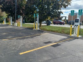 Uma área de estacionamento com estações de carregamento para veículos elétricos, vagas demarcadas e um edifício ao fundo.