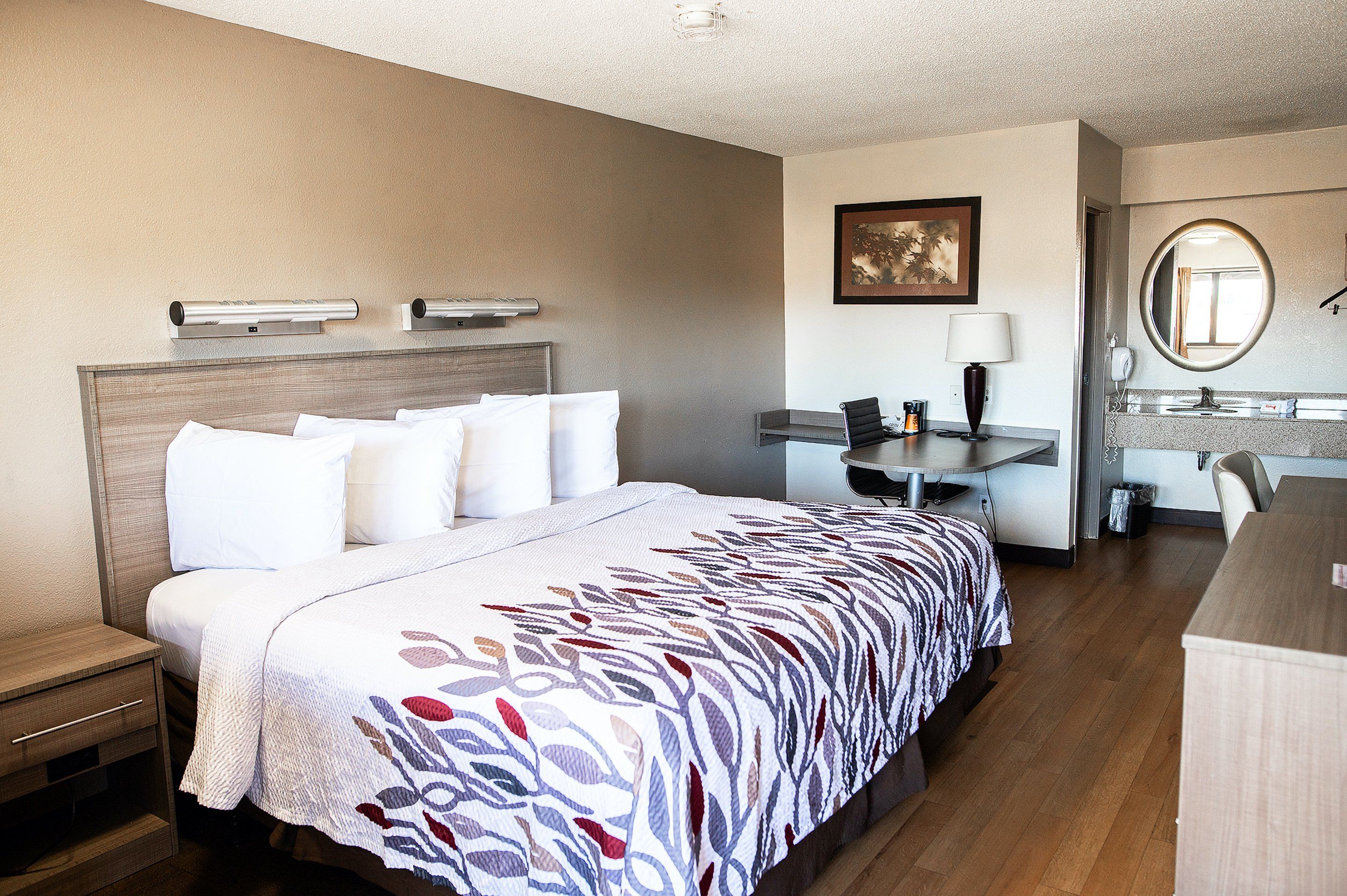 A hotel room with a bed, bedside table, desk, chair, and a mirror.
