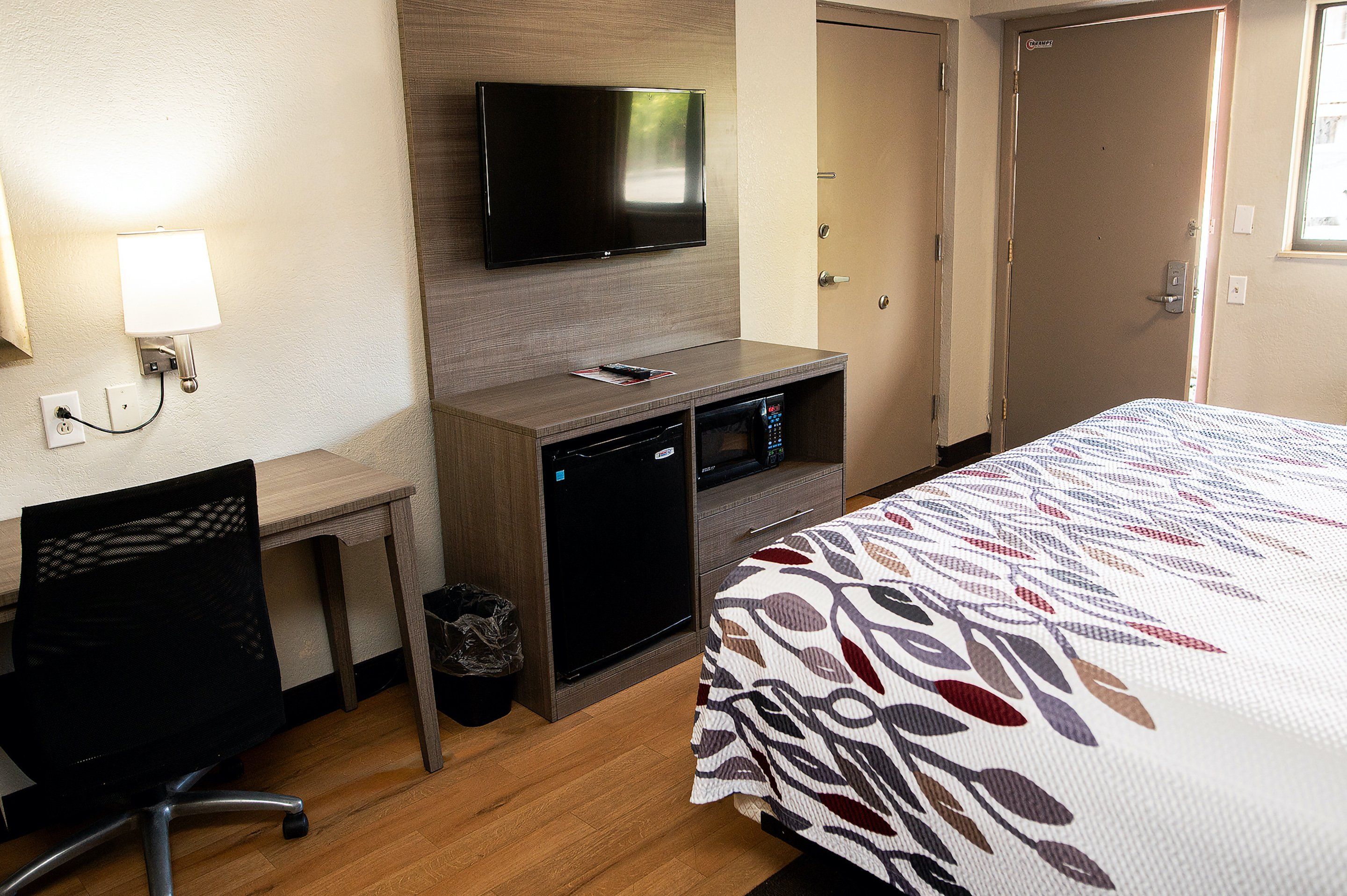 A hotel room with a bed, a desk, a television mounted on the wall, a microwave, and a trash can.