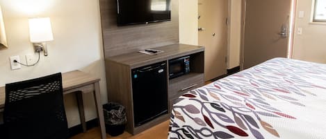 A hotel room with a bed, a desk, a television mounted on the wall, a microwave, and a trash can.