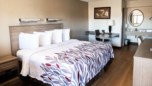 A hotel room with a bed, bedside table, desk, chair, and a bathroom with a mirror and sink. - Red Roof Inn Rochester - Henrietta (Henrietta)