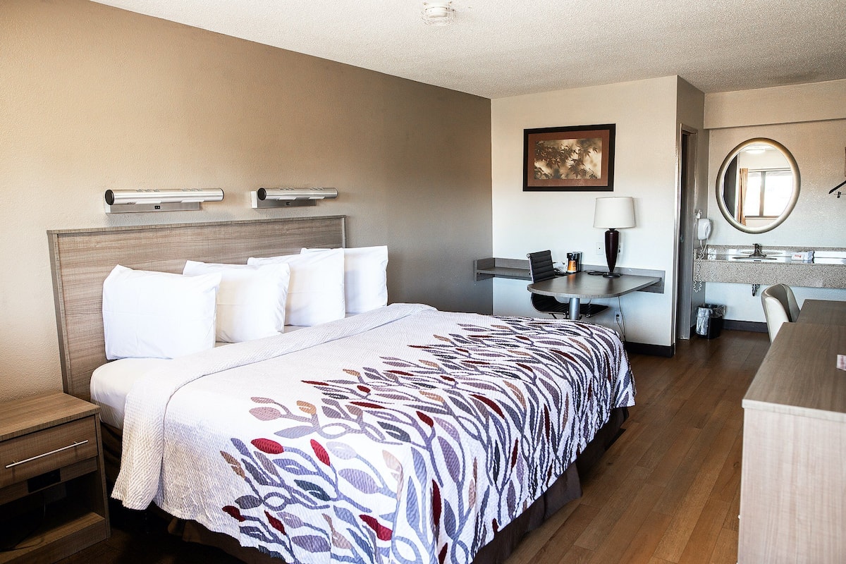 A hotel room with a bed, bedside table, desk, chair, and a bathroom with a mirror and sink.