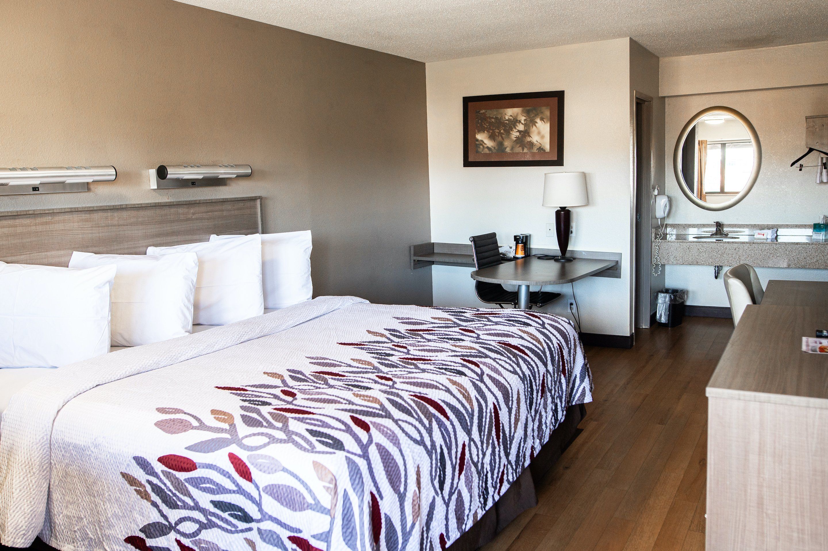 A hotel room with a bed, desk, chair, and a bathroom with a mirror and sink.