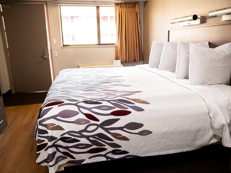 A hotel room with a bed, a dresser, a window with curtains, and a headboard with a light fixture.