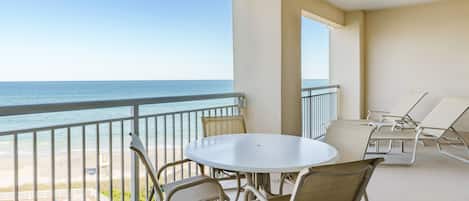 En balkon med hvidt bord og stole, med udsigt over en strand og hav.