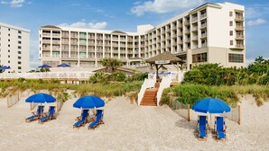Un hotel frente a la playa con varios balcones, una zona de playa con sillones y sombrillas, y una escalera que da a la playa.