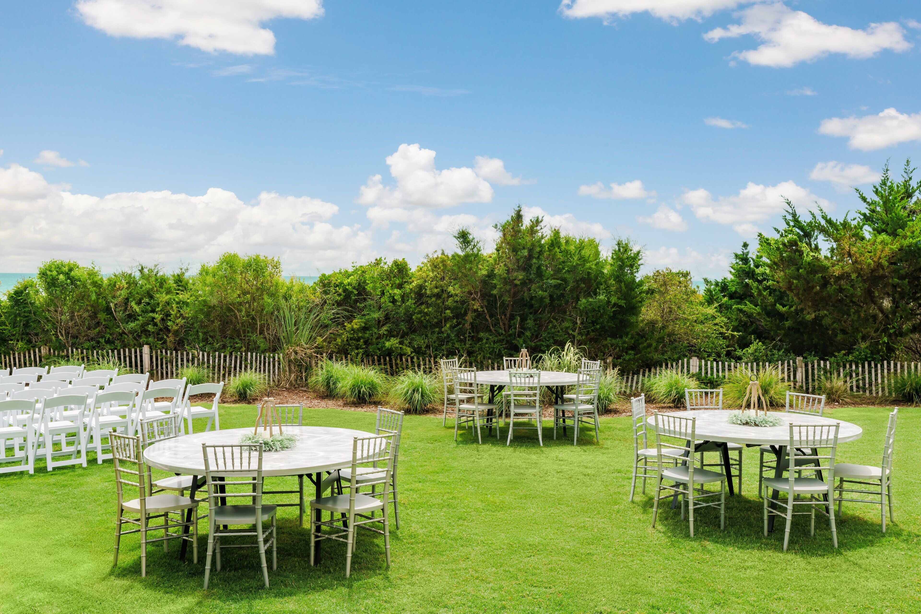 An outdoor setting with multiple round tables and chairs, surrounded by a wooden fence and lush greenery.