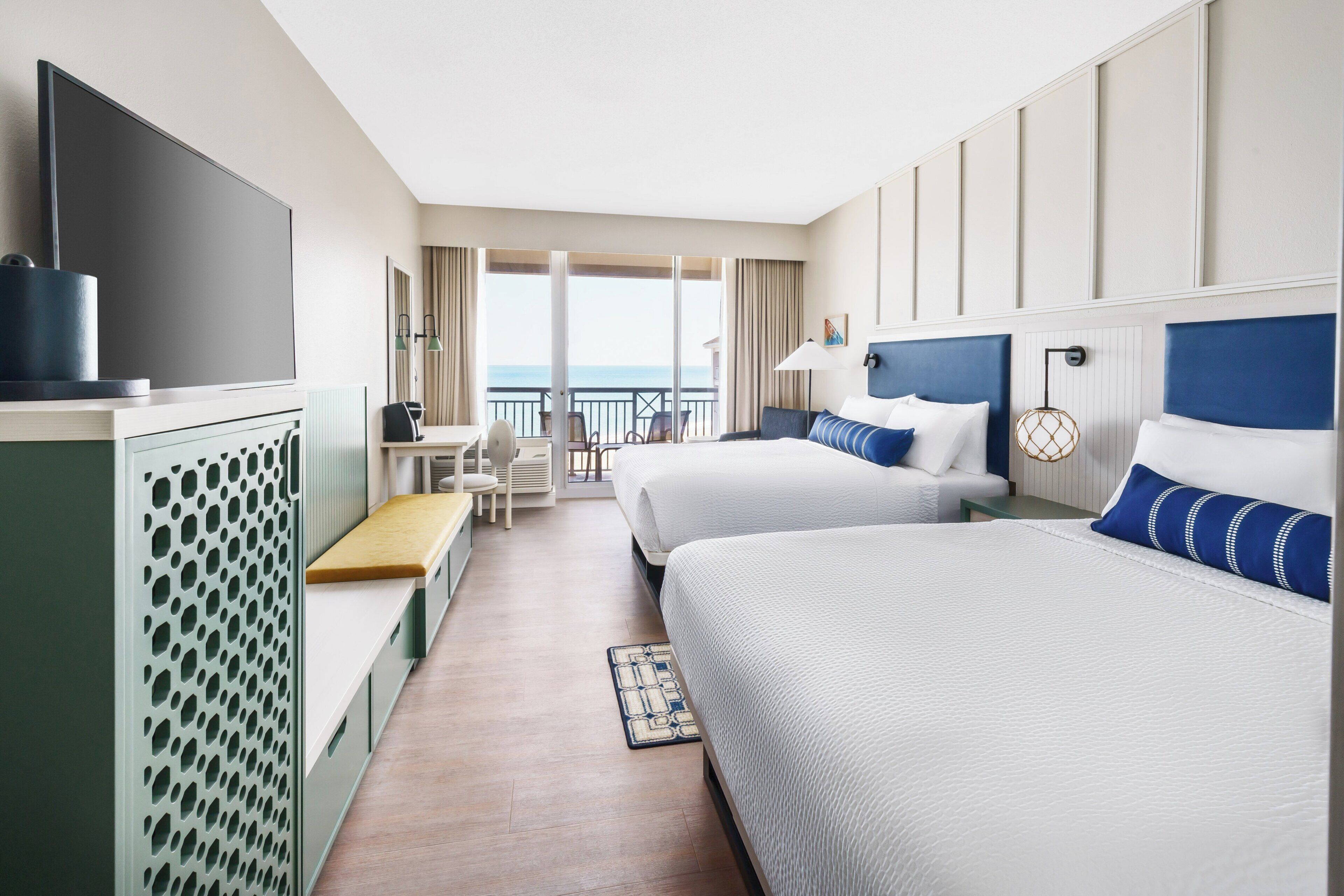 A hotel room with two beds, a desk, a chair, a TV, and a balcony with a view.