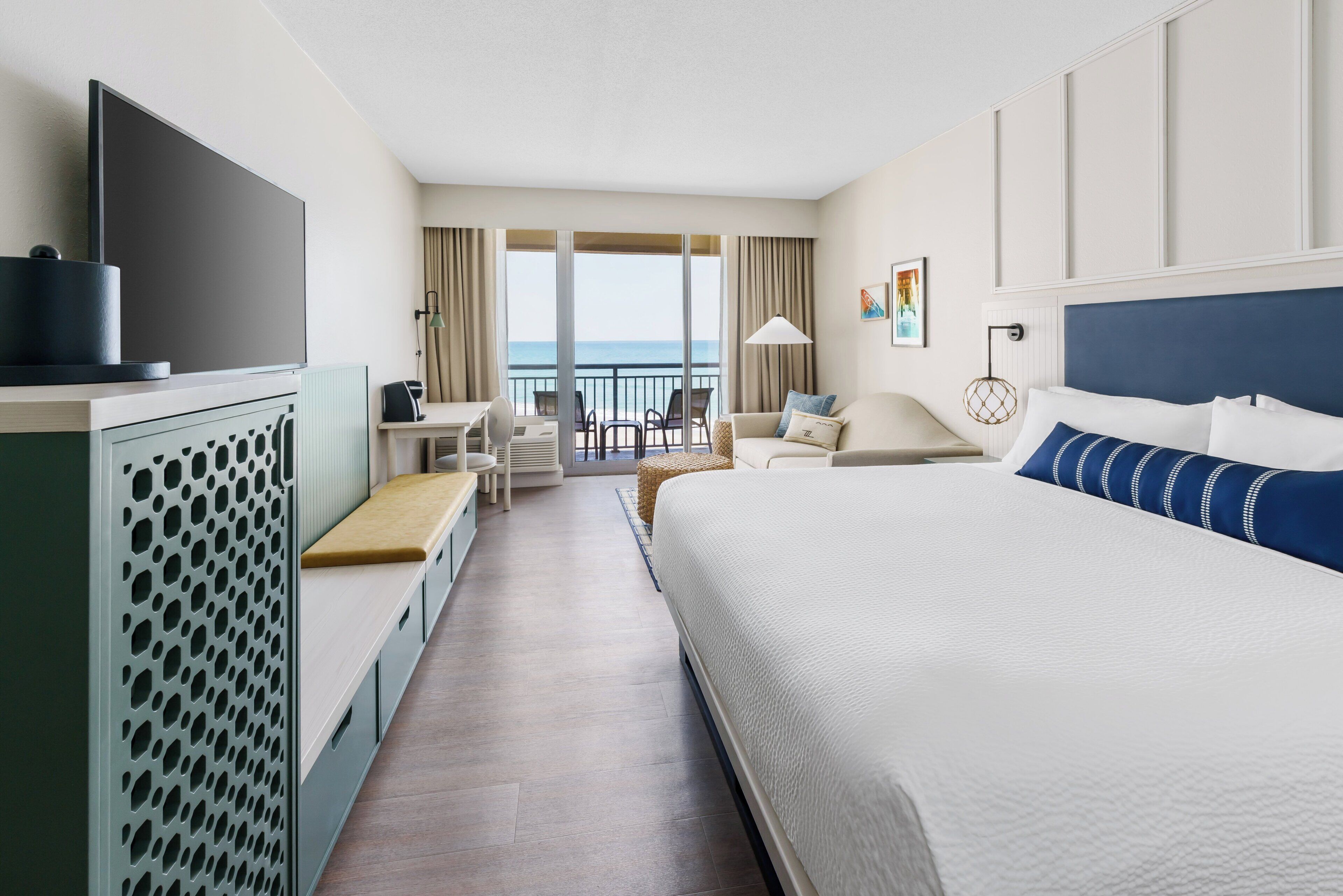 A hotel room with a large bed, a desk, a chair, a TV, and a view of the ocean.