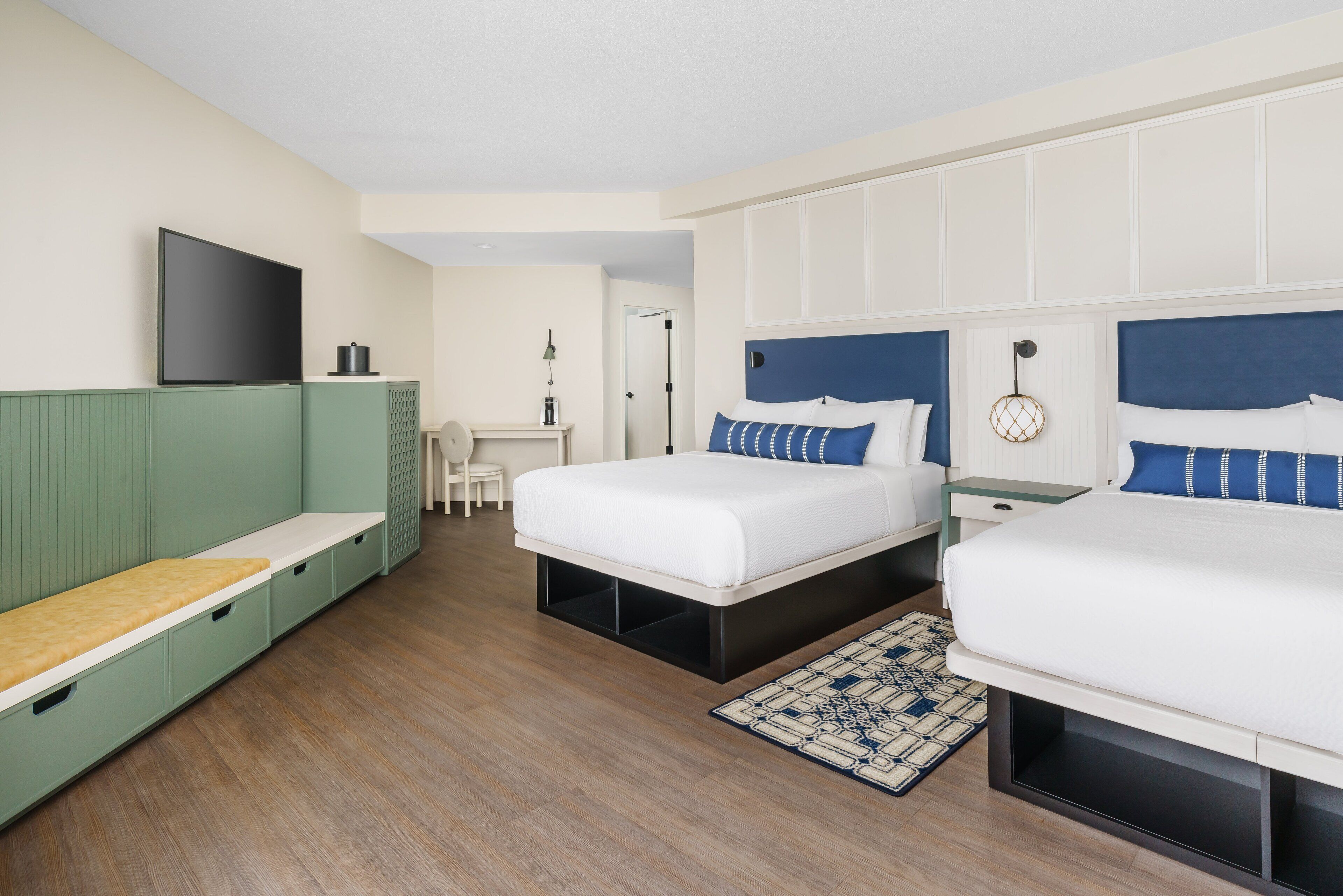 A hotel room with two beds, a TV, a desk, and a green bench.