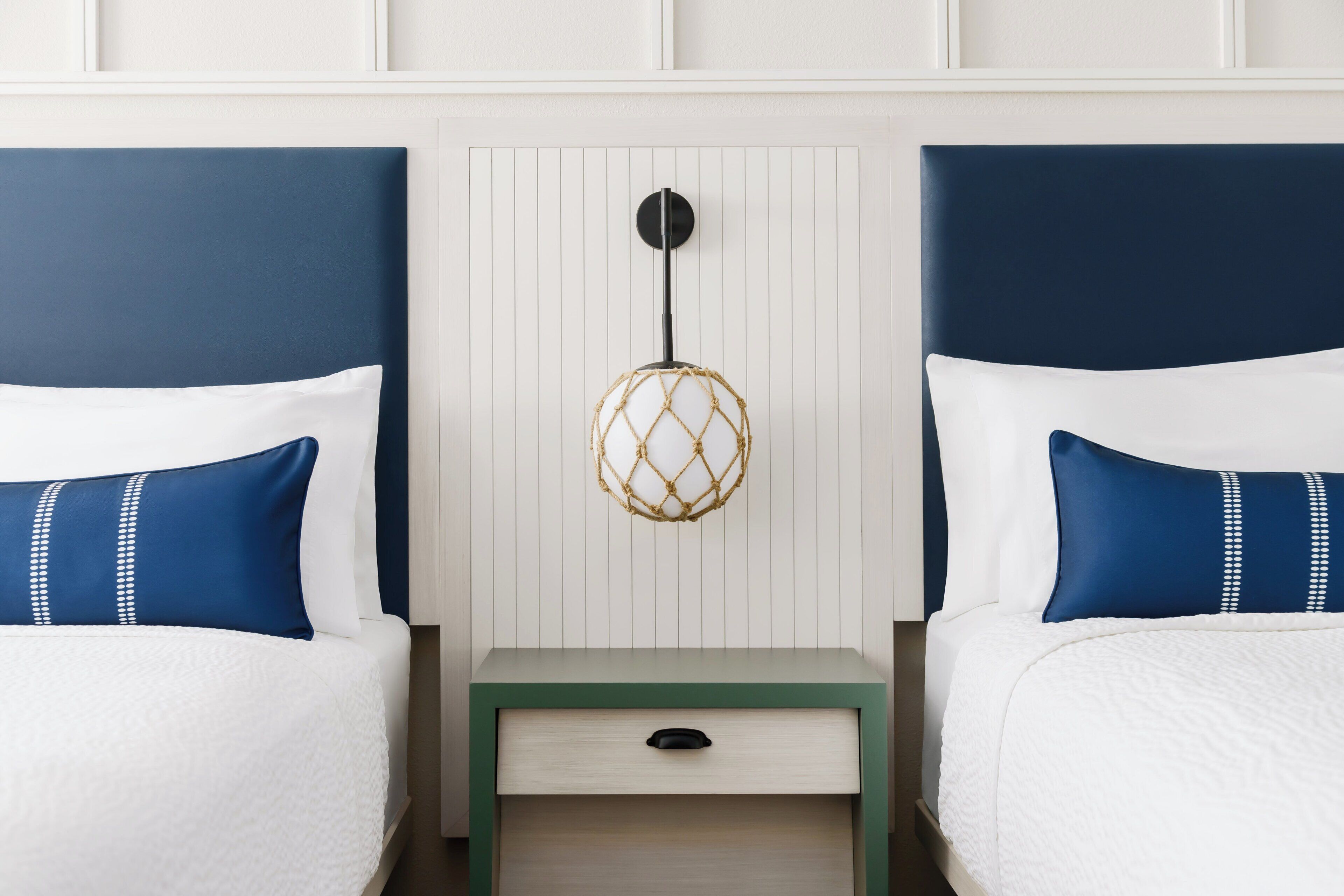 A double bed with white bedding and blue pillows with white dots, a small bedside table with a drawer, and a wall-mounted light fixture.