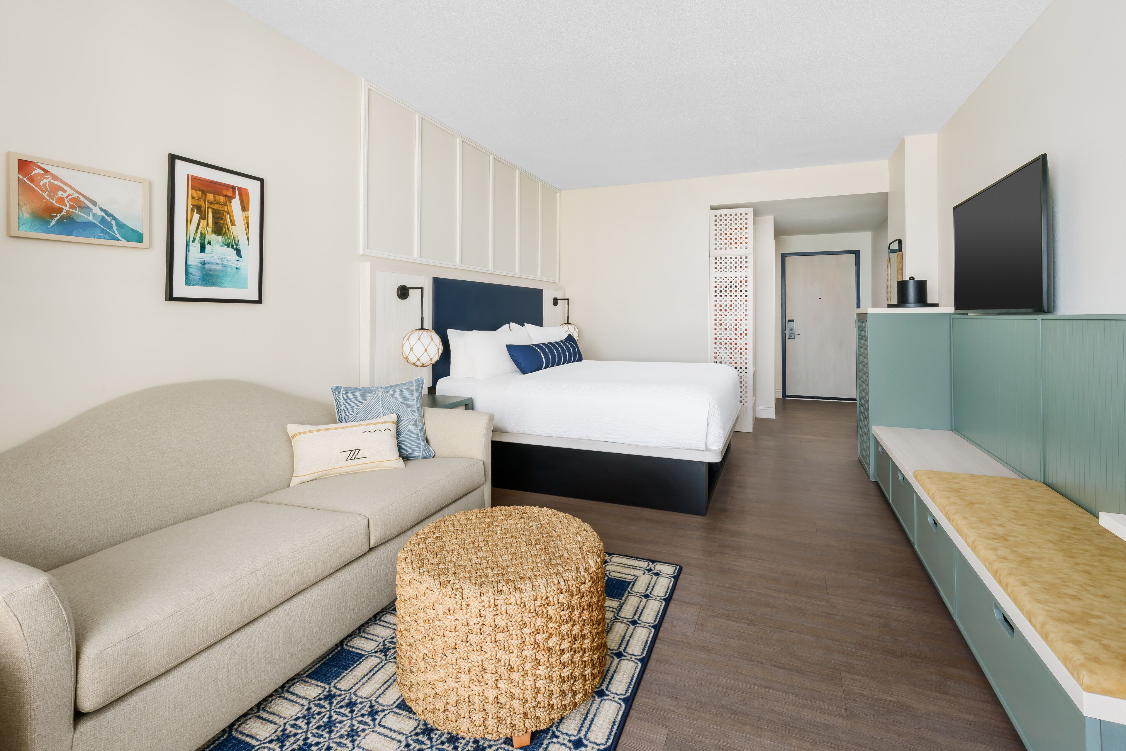Een moderne hotelkamer met een bed, een slaapbank, een rond poefje en een televisie op een standaard.