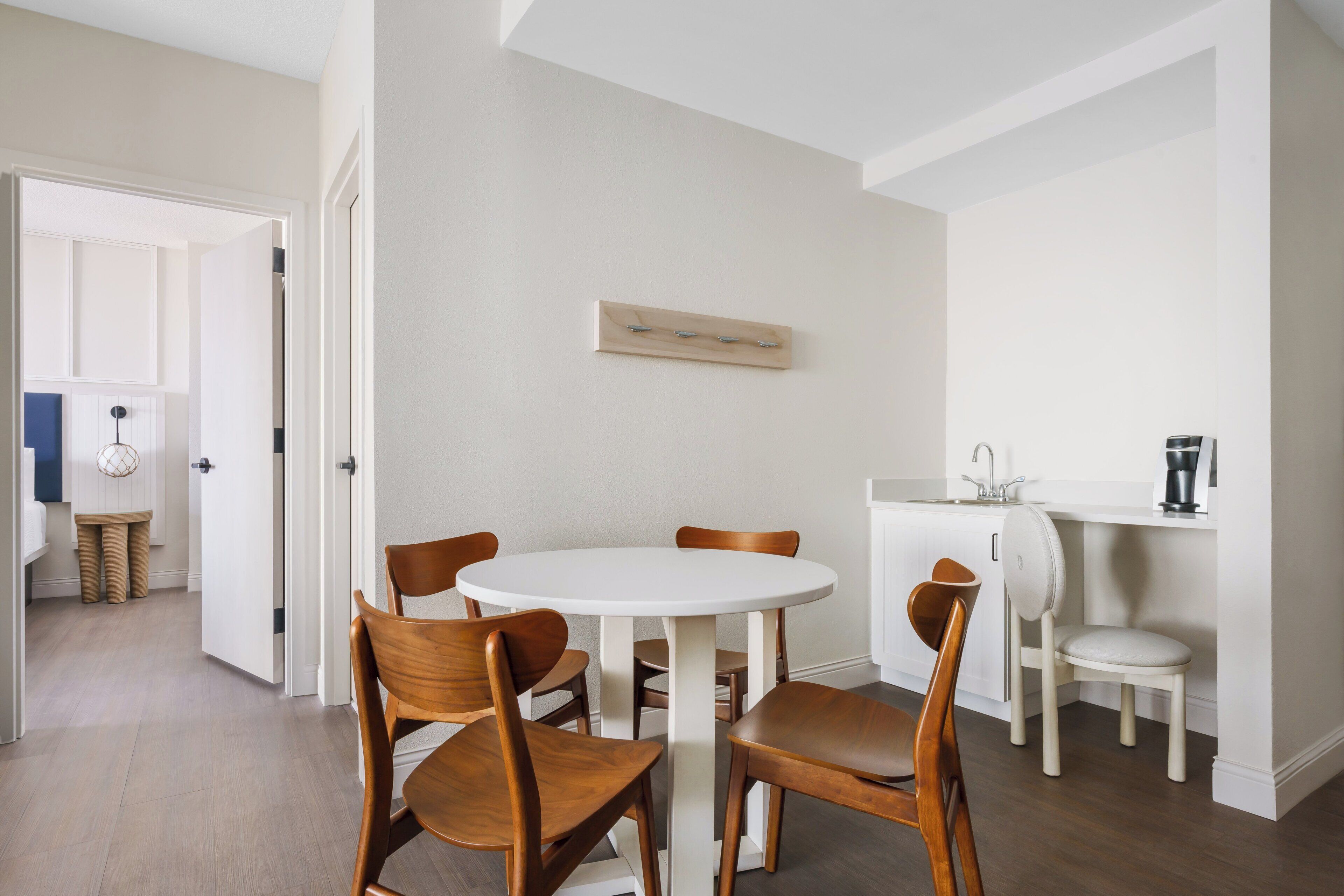 Een eetgedeelte met een ronde tafel en vier houten stoelen, een witte muur met een houten plank, en een keuken met een spoelbak en kastjes.