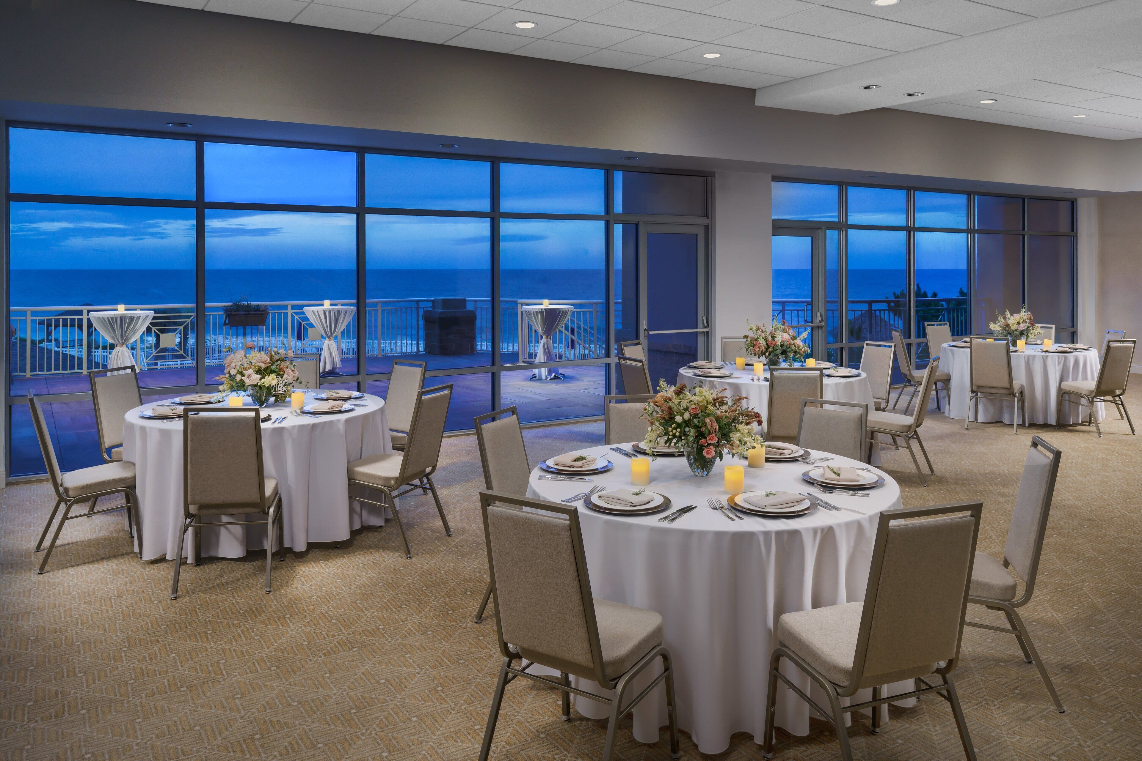 A dining area with round tables set for six, large windows offering a view of the ocean, and a balcony with tables and chairs.