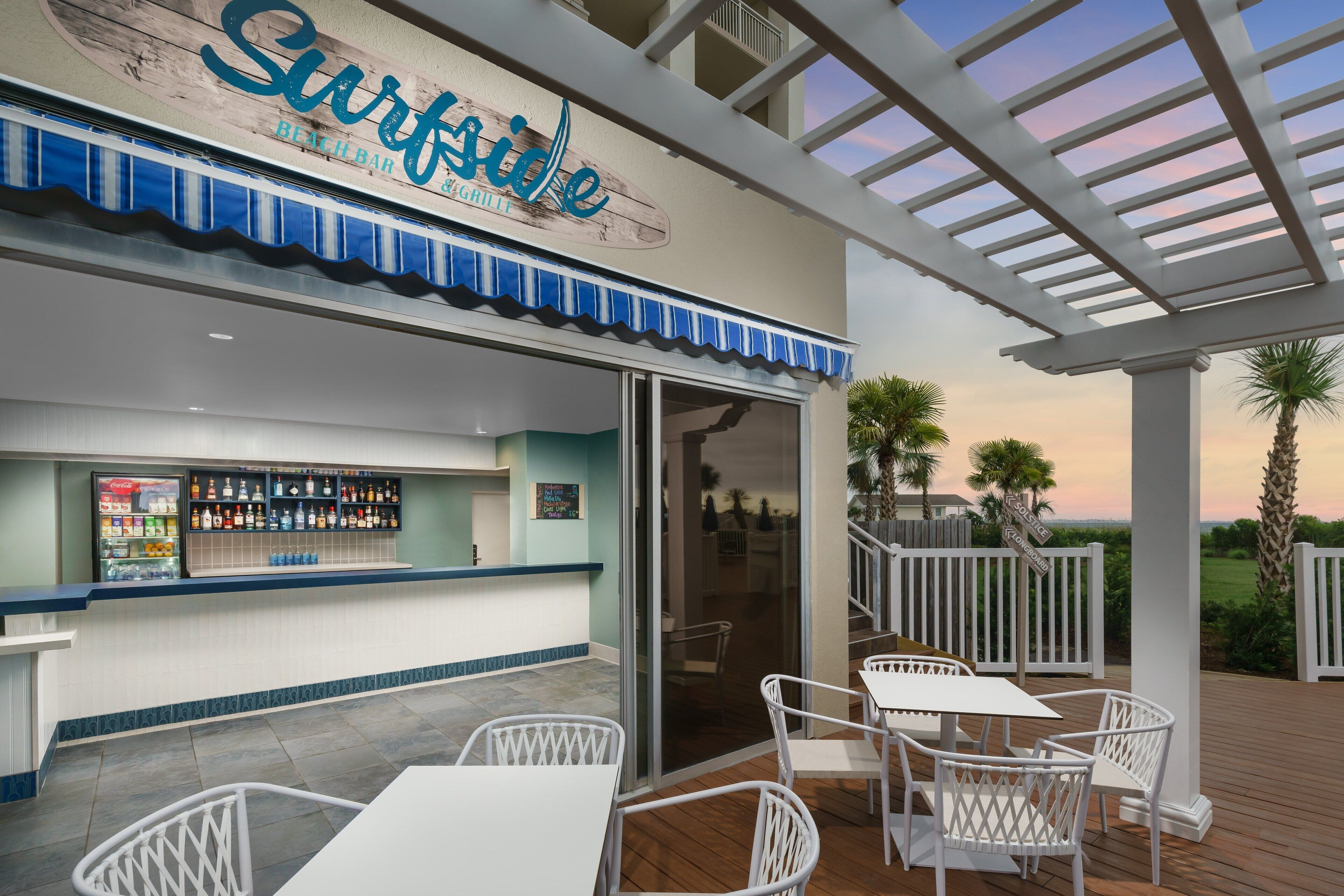 A beachside bar with a sign reading "Surfside Beach Bar & Grill," outdoor seating, and a view of the ocean.