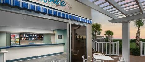 A beachside bar with a sign reading "Surfside Beach Bar & Grill," outdoor seating, and a view of the ocean.