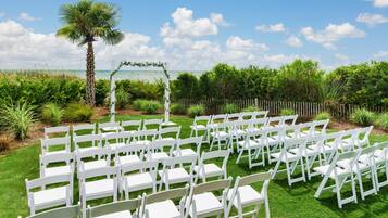 Une cérémonie de mariage en plein air, avec des chaises blanches disposées en rangées, un palmier et une clôture en bois en arrière-plan.