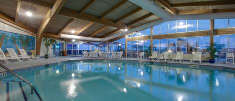 An indoor swimming pool with clear blue water, wooden beams on the ceiling, and white lounge chairs around the pool.