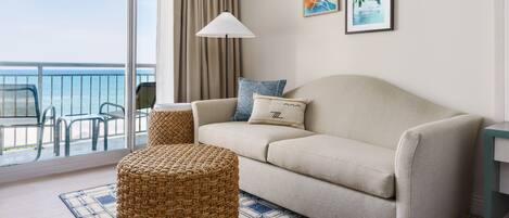 A modern living room with a beige sofa, a woven ottoman, and a patterned rug. There are two framed artworks on the wall, a floor lamp, and a view of the sea through large windows.