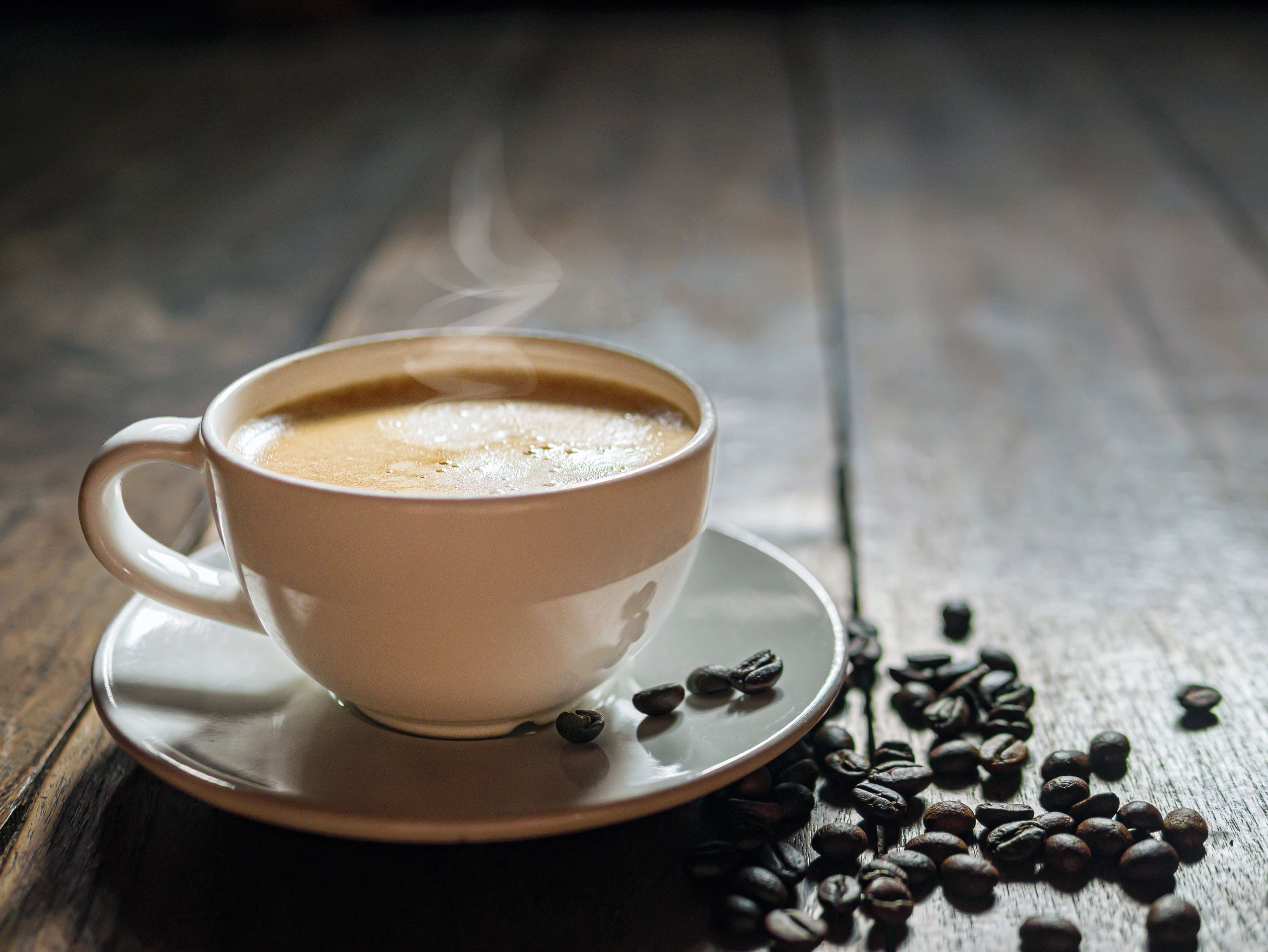 A white cup of coffee on a saucer with steam rising, surrounded by scattered coffee beans on a wooden surface.