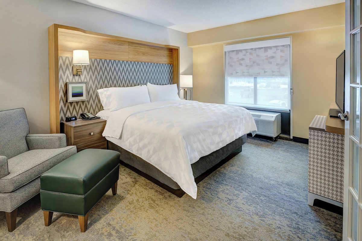 A hotel room with a bed, a chair, a bench, a bedside table with a lamp, a picture frame, and a window with a view.