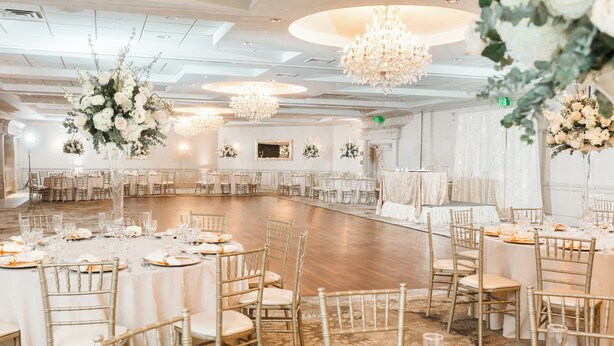 A large, elegantly decorated banquet hall with round tables, gold chairs, and hanging floral arrangements.
